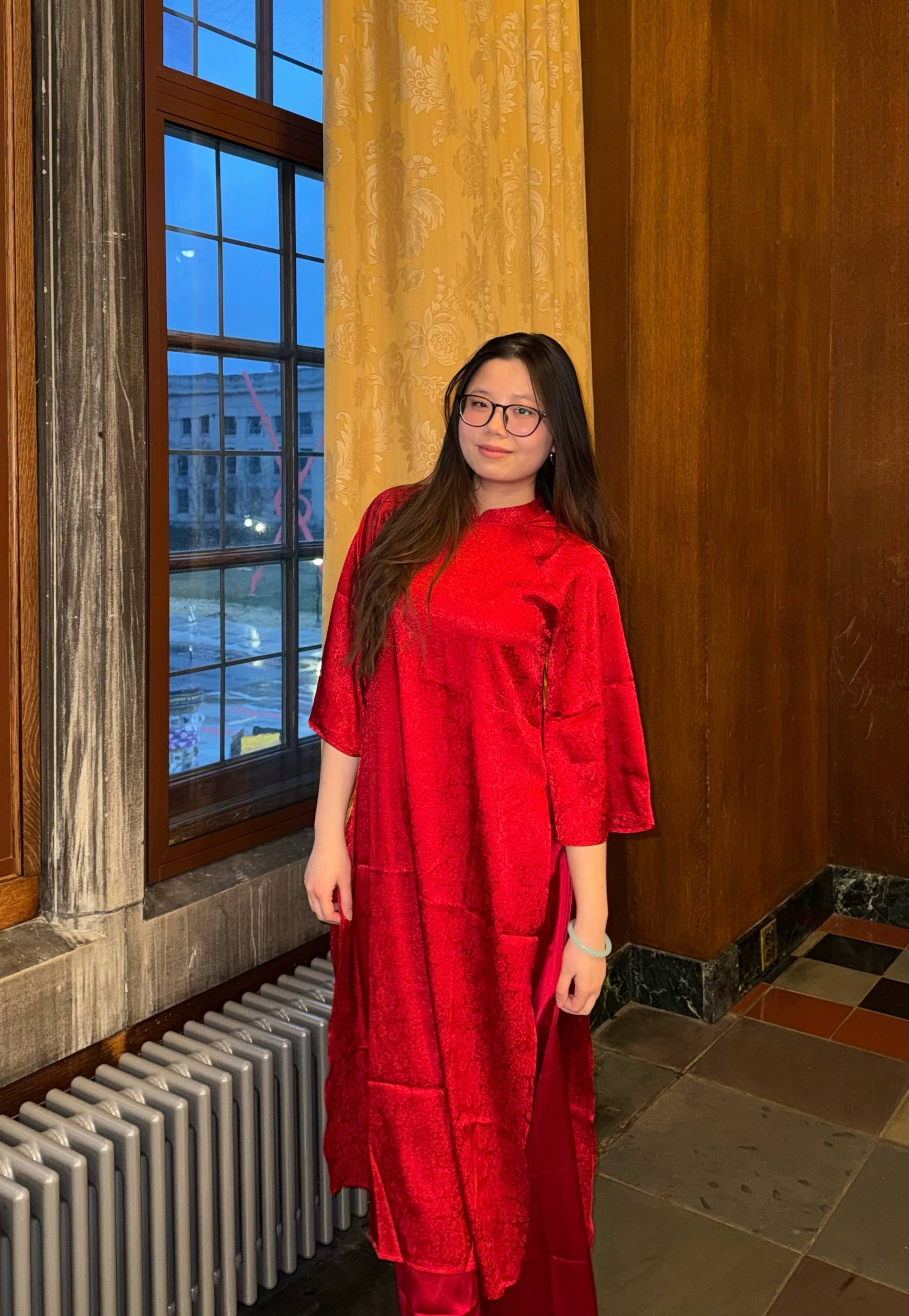 Student with long black hair and black glasses, wearing a red dress and standing in front of a window.